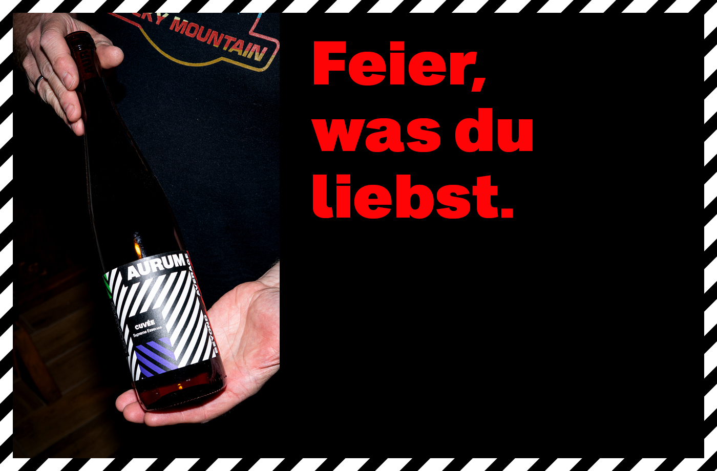 Plakat im markanten Schwarz mit diagonaler Rahmenstruktur. Rechts der rote Schriftzug: ‚Feier, was du liebst.‘ Links eine Hand, die eine Flasche ‚Aurum‘ mit grafisch schwarz-weiß gestreiftem Etikett präsentiert.“